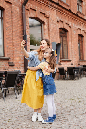 Full length portrait of happy mother and daughter with down syndrome taking selfie together outdoors in cityの写真素材