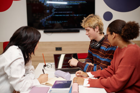 Diverse group of college students studying IT together at table in school library and using laptop with code lines on screenの写真素材