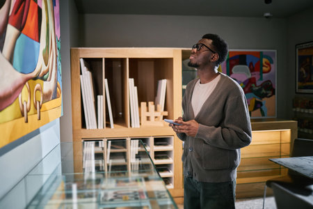 Person is viewing a colorful painting in modern museum setting, holding notes and showing interest in the artwork. Shelves with materials are visible in backgroundの写真素材