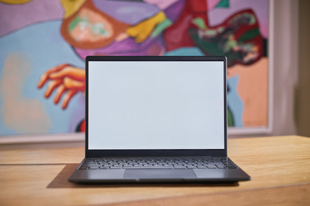 Modern laptop with blank screen placed on wooden desk in front of colorful abstract painting, ideal...