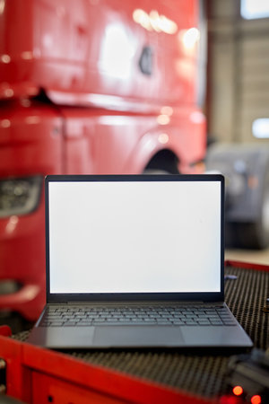 Laptop with blank screen situated on workbench in automotive garage. Red truck in blurred background and equipment surrounding the workspaceの写真素材