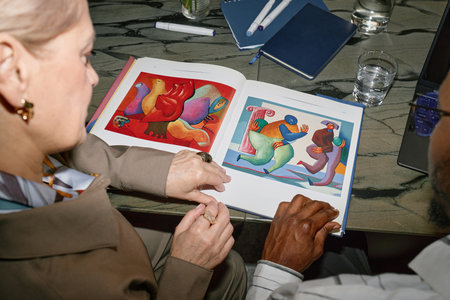 Two people closely examining colorful illustrations in an open book, seated at a table filled with notepads, pens, and glasses of water. Their hands pointing at the book pages with interestの写真素材