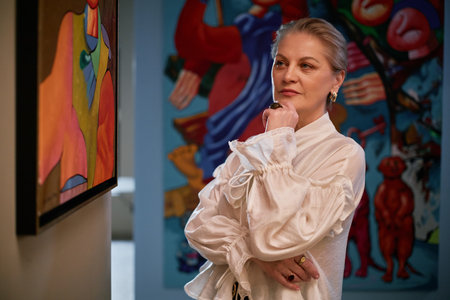 Woman observing abstract paintings in an art gallery during daytime, wearing elegant white blouse and thoughtful expression on face, with colorful artworks in background, exuding art enthusiast vibeの写真素材