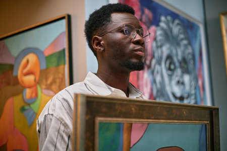 Portrait of African American man holding abstract painting in an art gallery. Showing concentration while examining details of framed artworkの写真素材