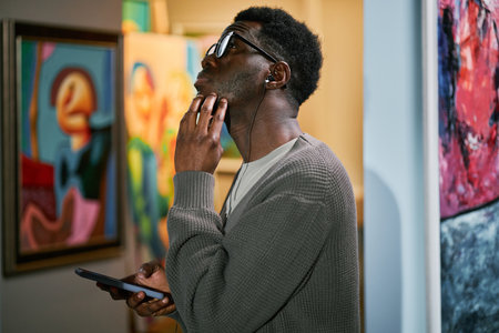 Man with headphones looking at painting in art gallery while holding phone. Engaging in thoughtful contemplation while surrounded by vibrant artwork in galleryの写真素材