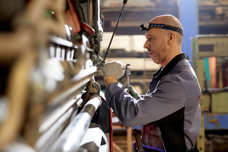 Technician wearing grey coveralls and headlight repairing complex industrial machinery seen from the side in a well-lit workshop. Metal wiring and various controls are visibleの写真素材
