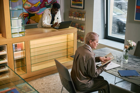 Mature woman sitting at desk during work meeting, writing in notepad. A man using a laptop computer visible in background, adding a modern, professional atmosphereの写真素材