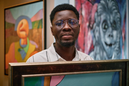 Portrait of an African American artist holding framed abstract artwork, standing in art gallery surrounded by colorful paintings. Glasses-wearing artist showcasing creative piecesの写真素材