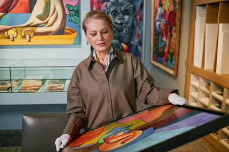 Senior woman holding and examining vibrant abstract artwork in an art gallery, with various other paintings visible in the backgroundの写真素材