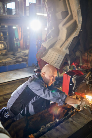 Bald mechanic working on vehicle engine in well-lit auto workshop environment. Wearing gray uniform and focused on his task, highlighting attention to detail and precisionの写真素材