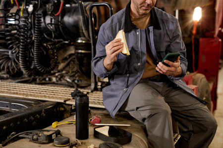 Mechanic sitting in workshop taking break by eating sandwich and using phone. Surrounded by various tools and equipment in industrial settingの写真素材
