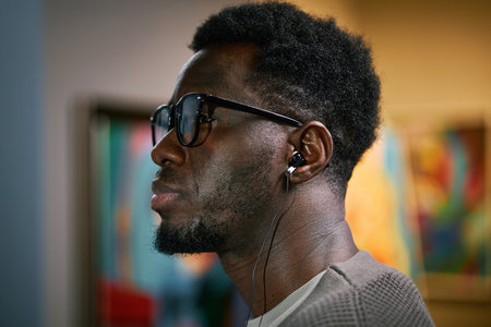 African American man enjoying music with earbuds while wearing sunglasses, standing in well-lit room with colorful abstract art in backgroundの写真素材