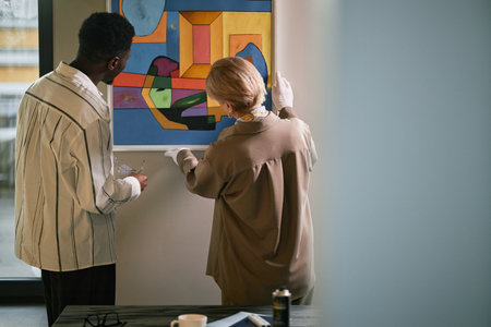 Two people engaged in closely examining vibrant modern art piece in gallery setting. Artwork featuring geometric shapes on vibrant colored backgroundの写真素材