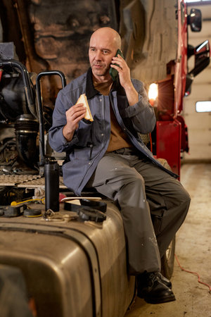 Mechanic in workshop speaking on phone while holding sandwich. Sitting on equipment, engaging in...