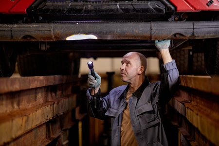 Mechanic examining car undercarriage in auto repair shop with flashlight, looking thoughtfully and focused on task at hand. Wearing work uniform while conducting inspectionの写真素材
