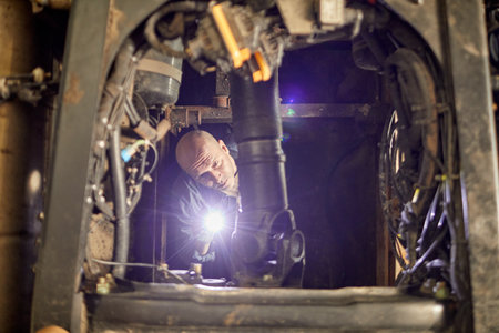 Mechanic repairing engine underneath machinery with intense focus and precision, highlighting intricate details of mechanical components. Man using flashlight for enhanced visibilityの写真素材