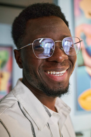 Portrait of smiling African American man wearing glasses while standing indoors. Background includes blurry colorful artworks hanging on the wallの写真素材