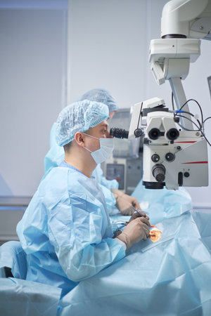 Middle aged man wearing surgical mask and cap performing surgery using operating microscope in...