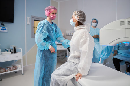 woman sitting on operating table wearing surgical gown and cap, female medical professional standing beside her preparing for procedure in operating roomの写真素材