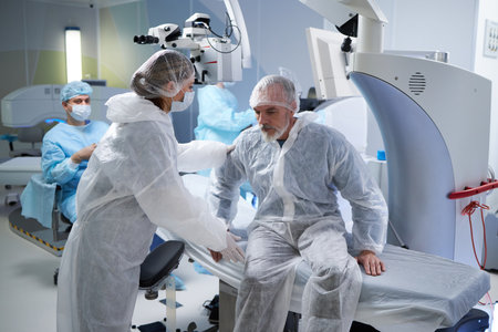 Middle aged man sitting on operating table assisted by woman in surgical gown while medical team...
