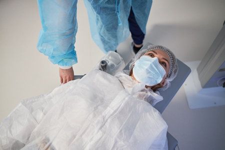 woman lying on operating table wearing surgical mask and cap, medical professional standing nearby...