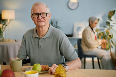Portrait of elderly man sitting at breakfast table enjoying healthy meal with another person in background appearing to knit. Smiling elderly man wearing glassesの写真素材