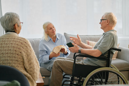 Senior man in wheelchair conversing with senior women in cozy living room setting, creating a warm and engaging atmosphere for social interactionの写真素材