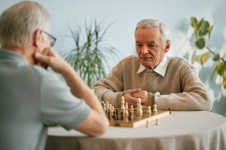 Senior men playing chess at indoor table. One man looking thoughtfully at chessboard while other man...