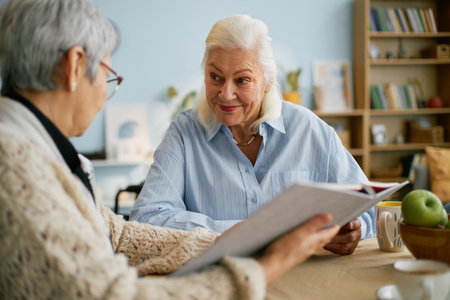 Elderly women sitting at table sharing moment while reading book in cozy setting. Warm atmosphere with bookshelves and houseplants surrounding them in backgroundの写真素材