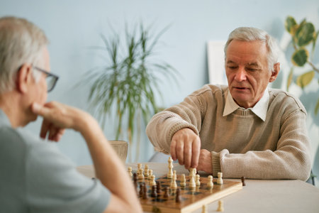Elderly man contemplating chess move against other senior player in a well-lit room with plant in backgroundの写真素材