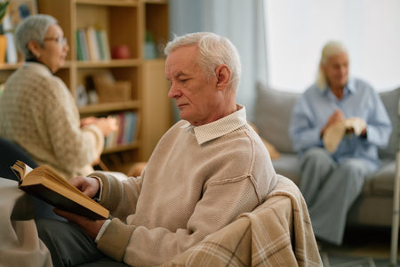 Elderly man engrossed in reading book while seated in a comfortable living room with other senior...