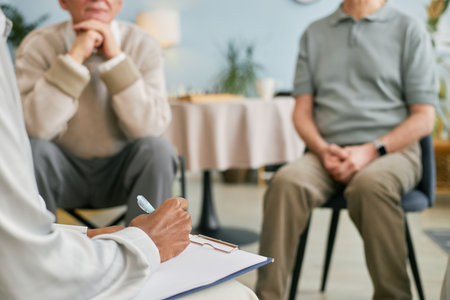 Seniors participating in group therapy session in comfortable medical office environment with therapist taking notes. Some participants appear thoughtful and engaged in conversationの写真素材