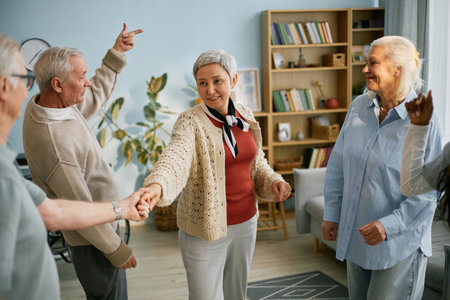 Seniors engaging in a cheerful group activity in a cozy living room, laughing and holding hands. Houseplants and bookshelves in background adding to the homely atmosphereの写真素材