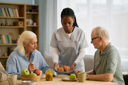 Elderly couple sitting at table having breakfast while caregiver bringing pastries on plateの写真素材