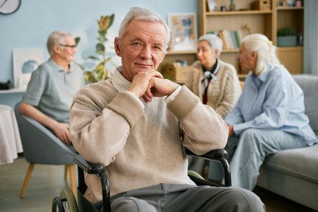 Senior man in wheelchair posing in living room with other elderly individuals in background engaging in conversation. Warm and cozy ambiance with natural daylightの写真素材