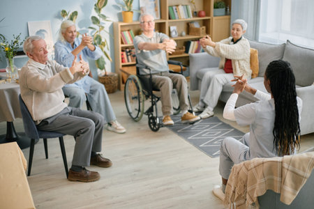 Group of elderly individuals participating in seated exercise session led by an instructor in a cozy room social interaction and physical healthの写真素材
