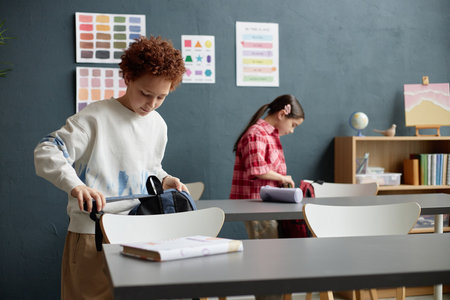 Caucasian boy and Caucasian girl standing in classroom packing backpacks on desks, focusing on...