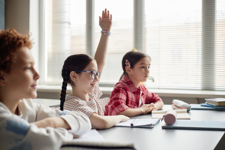 Three children sitting at classroom desk, Caucasian girl with glasses raising hand while smiling,...