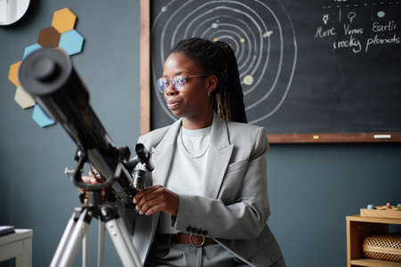 Young adult Black woman adjusting telescope in classroom with solar system diagram on chalkboard in...