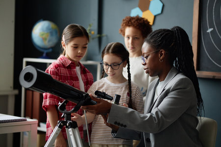 Black woman teacher demonstrating telescope to three children including two girls and one boy in classroom setting, with globe and chalkboard visible in backgroundの写真素材
