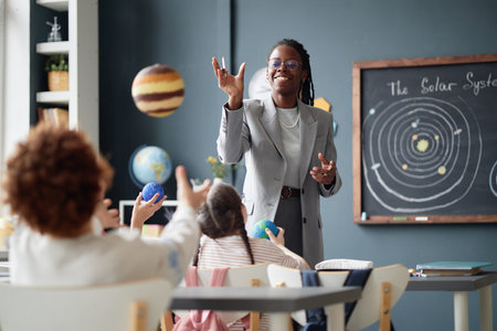 Black woman teacher engaging group of children in science classroom, tossing model planets while kids reaching to catch them, solar system diagram visible on chalkboard in backgroundの写真素材