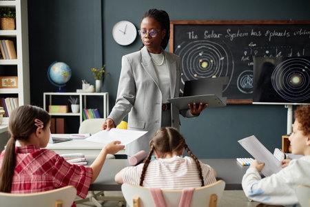 Black woman teacher handing worksheets to group of children sitting at classroom table, children...