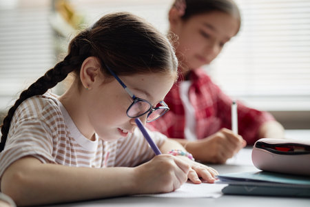 girl with glasses writing in notebook, sitting at desk with another girl in background studying, both appearing as children focused on schoolwork in classroom settingの写真素材
