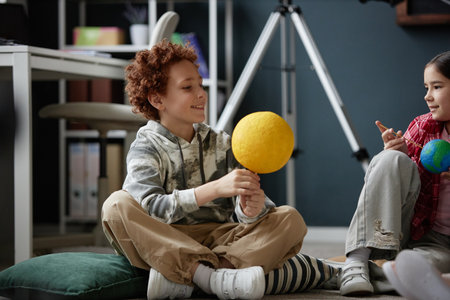 Caucasian boy and girl sitting on floor holding model planets, smiling and interacting during...