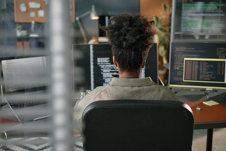 Back view of young Black woman sitting at desk working on computer with multiple monitors displaying...
