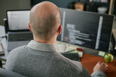 Middle aged Caucasian man sitting at desk working on computer monitors with programming code visible...