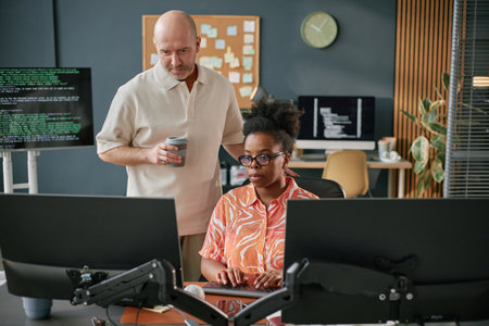 Black young adult woman typing on computer keyboard while Caucasian middle aged man standing beside...