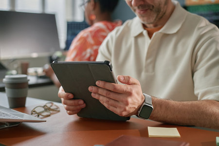 Middle aged Caucasian man using digital tablet at desk in modern office, sitting near Black woman...