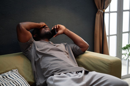 Black man sitting on sofa holding smartphone to ear with one hand covering face appearing stressed, indoor scene suggesting house arrest situation with visible window in backgroundの写真素材