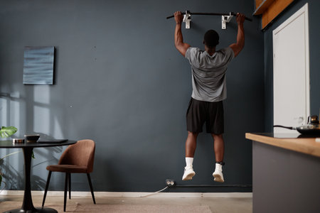 Black young adult man performing pull up exercise on wall mounted bar in modern apartment,...
